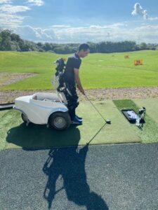 MAN IN PARAGOLFER MACHINE STANDING UP HITTING GOLF BALL WITH IRON